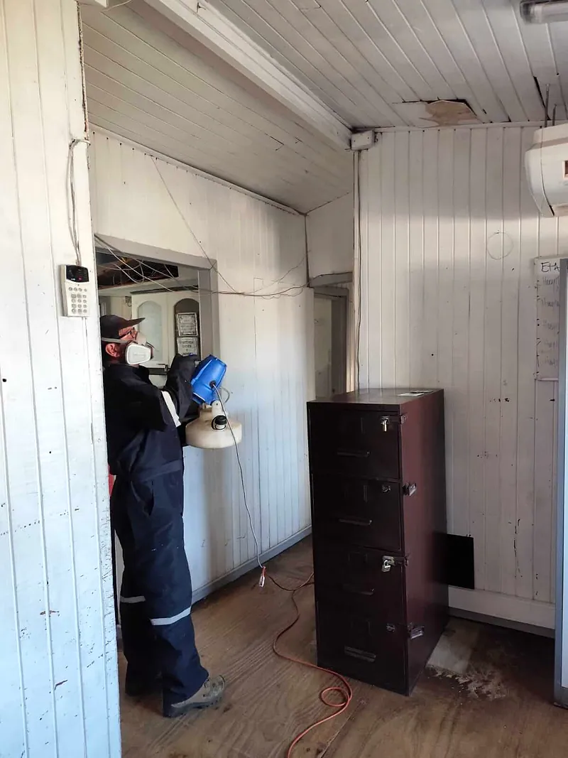 Técnico con máscara de doble filtro nebuliza el pasillo de una oficina con un equipo nebulizador eléctrico