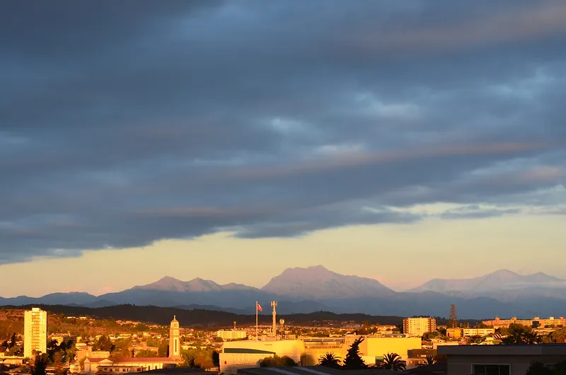 Quilpué al atardecer: edificios y una torre iluminados, con el cerro La Campana al fondo